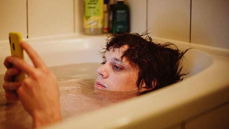 Zeno (Simon Steinhorst) in der Badewanne und scrollt durch sein Handy auf der Suche nach Kontakten. (Foto: SWR, Trimafilm)