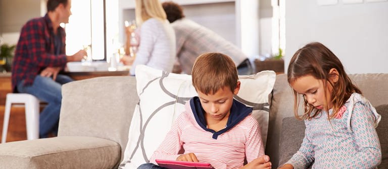 Ein Junge und ein Mädchen mit Smartphone und Tablet auf dem Sofa, im Hintergrund Erwachsene (Foto: Getty Images)