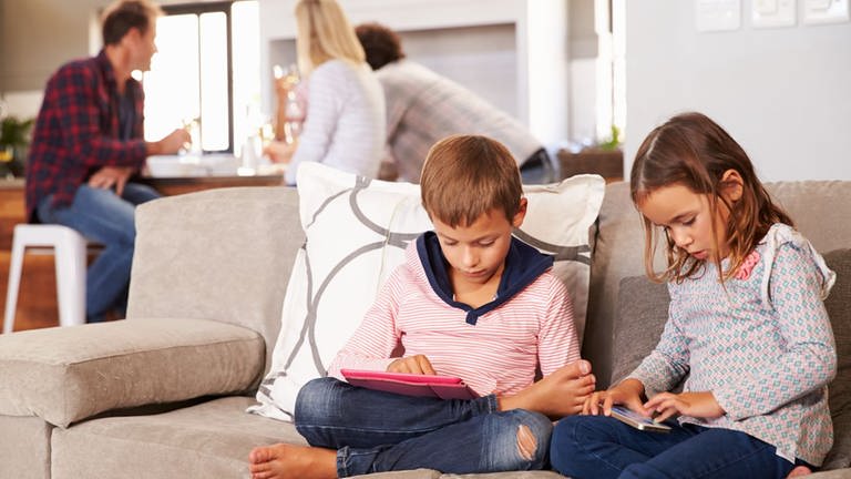 Ein Junge und ein Mädchen mit Smartphone und Tablet auf dem Sofa, im Hintergrund Erwachsene (Foto: Getty Images)
