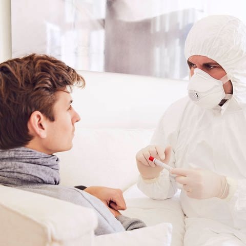 Arzt in Schutzkleidung untersucht einen Patienten zu Hause (Foto: IMAGO, imago images / Westend61)