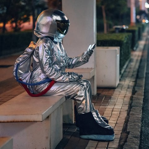 Eine als Astronaut verkleidete Person sitzt nachts an einer Bushaltestelle (Foto: imago images, IMAGO / Westend61)