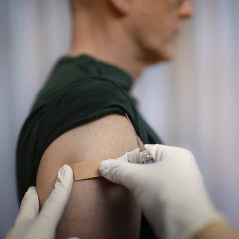 Arzt klebt nach der Impfung Pflaster auf den Arm eines Patienten: Manche Menschen spüren nach der Impfung keinerlei Nebenwirkung; das Immunsystem wurde dennoch aktiviert (Foto: IMAGO, IMAGO / photothek)