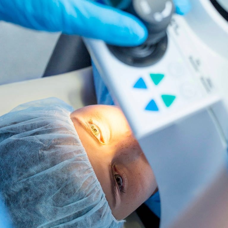 Operation am Auge einer Patienten mithilfe der Lasik-Methode. Die liegende Patientin trägt eine OP-Haube, ihr linkes Auge wird von einem medizinischen Gerät über ihr angeleuchtet. (Foto: IMAGO, IMAGO / Science Photo Library)