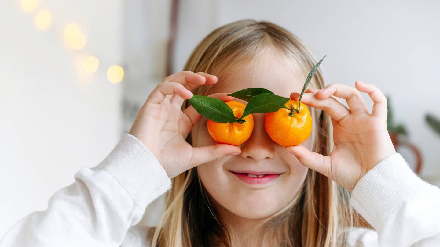 Ein blondes Mädchen hält sich lächelnd zwei Mandarinen vor die Augen: Eine gesunde, vitaminreiche Ernährung ist wichtig für die Augen (Foto: IMAGO, IMAGO / Westend61)