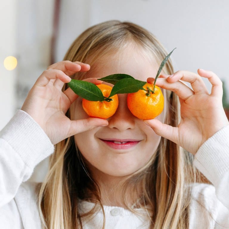 Ein blondes Mädchen hält sich lächelnd zwei Mandarinen vor die Augen: Eine gesunde, vitaminreiche Ernährung ist wichtig für die Augen (Foto: IMAGO, IMAGO / Westend61)