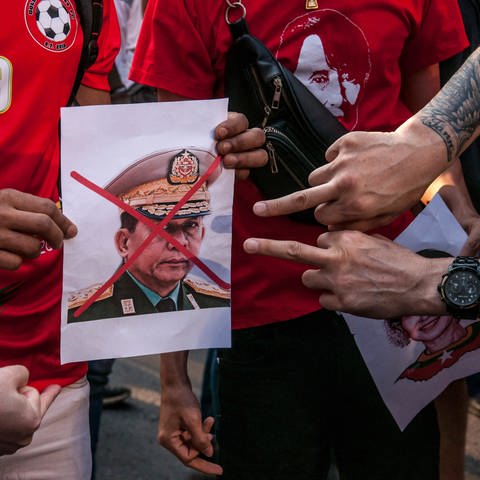 Demonstranten halten ein Porträt von Armeechef Min Aung Hlaing, über dessen Gesicht ein rotes Kreuz gemalt wurde. Eine zeigen mit dem Mittelfinger auf das Bild: Das Militär hatte am 1. Februar 2021 die zivile Führung Myanmars um Aung San Suu Kyi durch einen Putsch entmachtet und einen einjährigen Ausnahmezustand verhängt. Das Wort "Putsch" kommt ursprünglich aus der Schweiz. (Foto: dpa Bildfunk, picture alliance/dpa/SOPA Images via ZUMA Wire | Peerapon Boonyakiat)