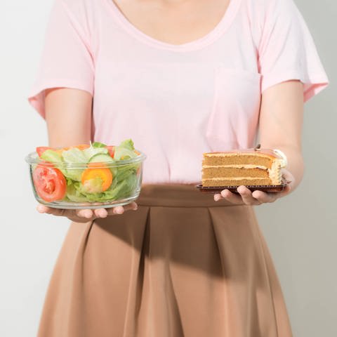 Frau hält in einer Hand eine Schale Salat, in der anderen ein Stück Torte: Eine Ernährungsumstellung ist ein langer Prozess. Man braucht für eine Veränderung des eigenen Essverhaltens Antrieb von außen, z.B. vom Hausarzt oder der Familie. Die kann motivieren und unterstützen: "Das hast du gut gemacht" oder "Komm, probier's noch mal".  (Foto: IMAGO, IMAGO / YAY Images)