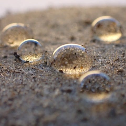 Wassertropfen sitzen wie Perlen auf der Oberfläche eines sandigen Bodens: Wenn es nach einer längeren Dürre plötzlich wieder stärker regnet, können die Böden nicht so viel Wasser auf einmal aufnehmen und es auch nicht speichern (Foto: © BGR)