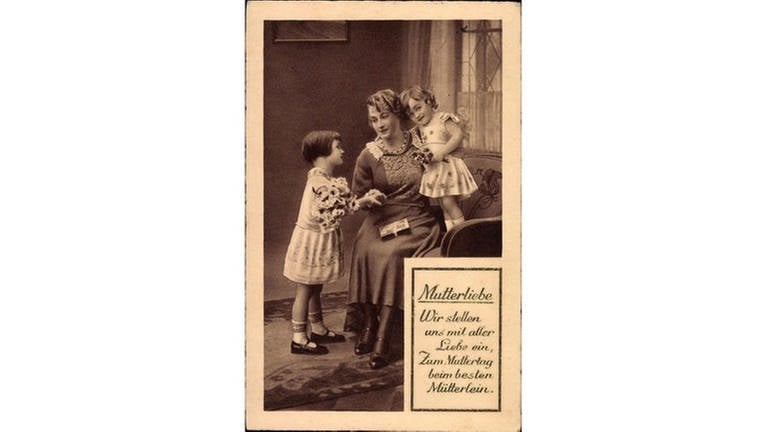 Mutter mit ihren zwei kleinen Töchtern, die ihr Blumen schenken (Foto): Glückwunschkarte zum Muttertag, um 1933, mit der Aufschrift "Mutterliebe – Wir stellen uns mit aller Liebe ein, zum Muttertag beim besten Mütterlein" (Foto: IMAGO, IMAGO / Arkivi)