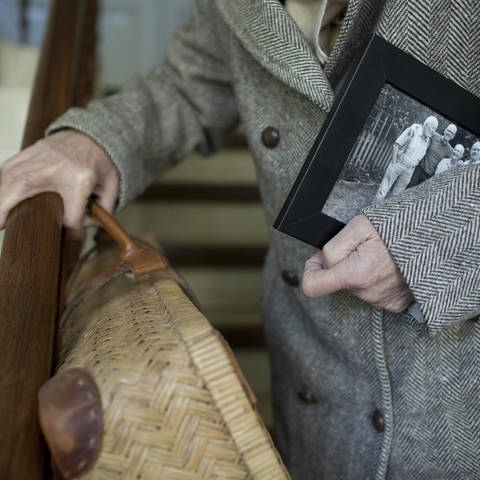 Seniorin mit Koffer und Familienfoto unter dem Arm steigt eine Treppe hinunter: Orientierungsstörungen gehören auf mit zur Demenz. Die sind am Anfang zwar nicht so deutlich, nehmen aber zu. (Foto: IMAGO, IMAGO / photothek)
