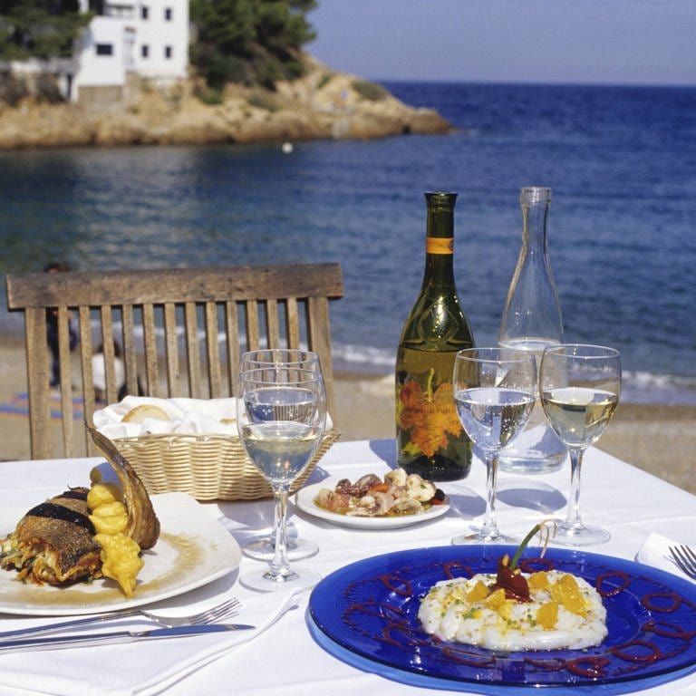 Gedeckter Tisch mit Wein am Strand: Man bringt Wein aus den Ferien mit, der vor Ort sehr lecker war, trinkt ihn zu Hause und wundert sich, weil er nicht schmeckt. Warum ist das so?  (Foto: IMAGO, IMAGO / Westend61)