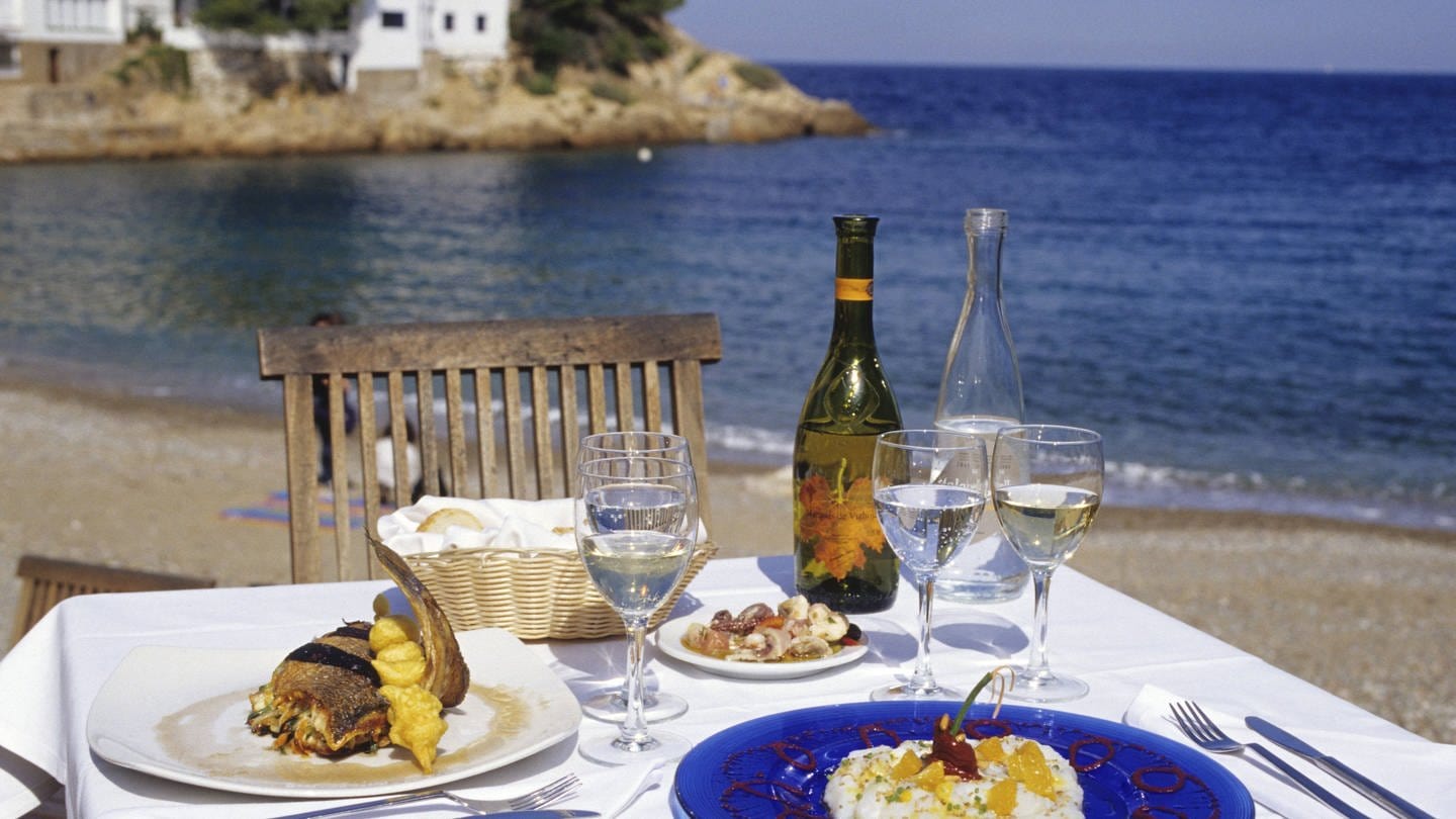 Gedeckter Tisch mit Wein am Strand: Man bringt Wein aus den Ferien mit, der vor Ort sehr lecker war, trinkt ihn zu Hause und wundert sich, weil er nicht schmeckt. Warum ist das so? (Foto: IMAGO, IMAGO / Westend61)