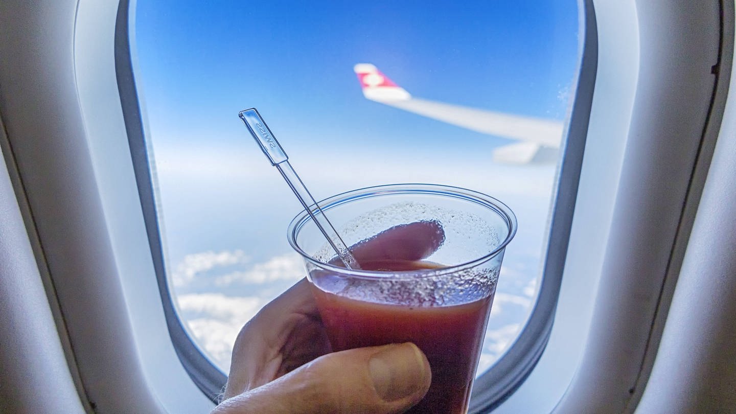 Eine Hand hält einen Becher mit Tomatensaft vor einem Fenster mit Blick aus einem Flieger: Der Geschmackssinn verändert sich im Flugzeug. Das wird beim Tomatensaft besonders deutlich. (Foto: IMAGO, IMAGO / Arnulf Hettrich)