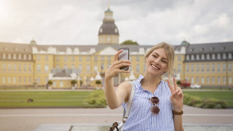 Junge Frau macht vor dem Schloss in Karlsruhe ein Selfie: Fotos von Gegenständen mit dem Handy werden uns so gezeigt, wie wir sie selbst sehen. Aber ein Selfie ist immer spiegelverkehrt. Wie kommt das? (Foto: IMAGO, IMAGO / Westend61)