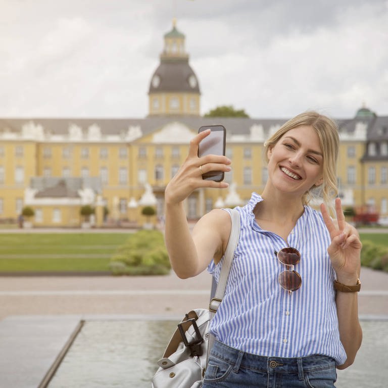 Junge Frau macht vor dem Schloss in Karlsruhe ein Selfie: Fotos von Gegenständen mit dem Handy werden uns so gezeigt, wie wir sie selbst sehen. Aber ein Selfie ist immer spiegelverkehrt. Wie kommt das? (Foto: IMAGO, IMAGO / Westend61)