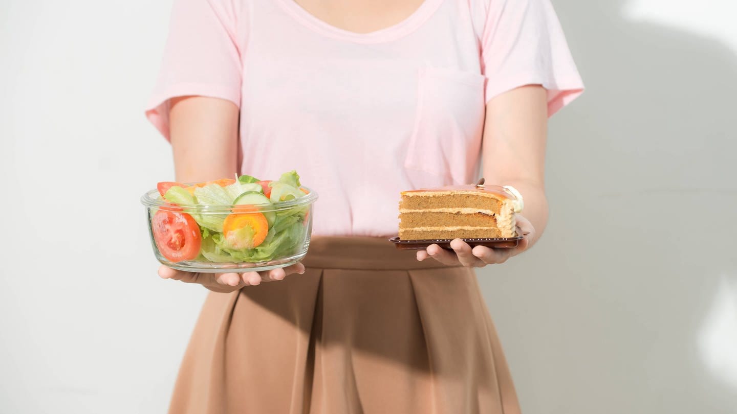 Frau hält in einer Hand eine Schale Salat, in der anderen ein Stück Torte: Eine Ernährungsumstellung ist ein langer Prozess. Man braucht für eine Veränderung des eigenen Essverhaltens Antrieb von außen, z.B. vom Hausarzt oder der Familie. Die kann motivieren und unterstützen: 