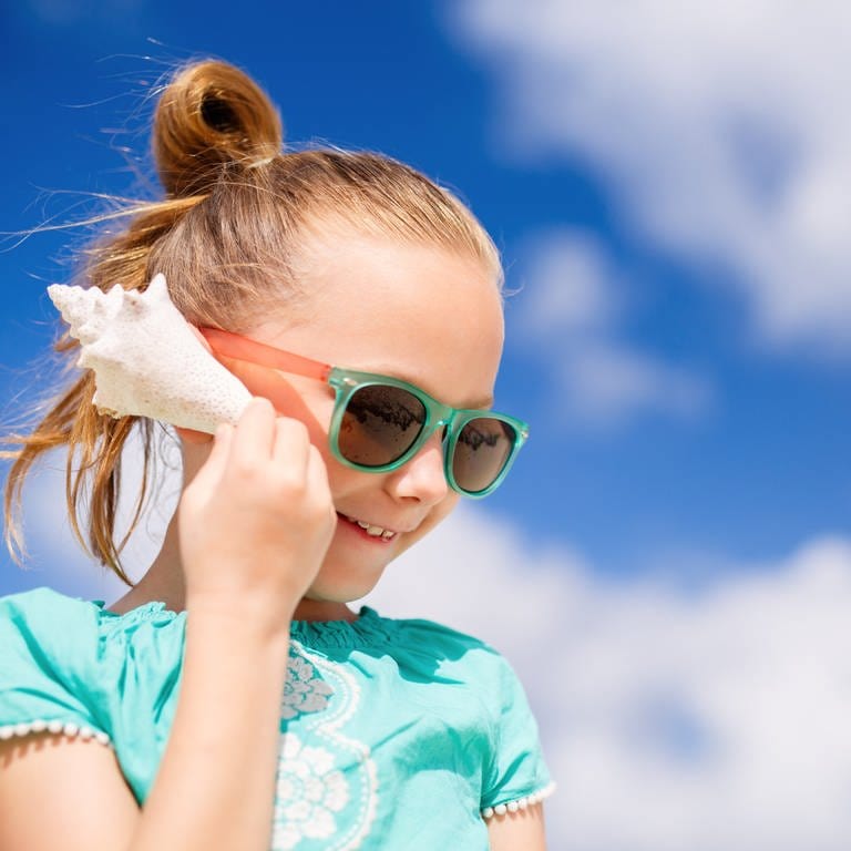 Ein Mädchen hält sich eine Meeresschnecke ans Ohr: Was wir in Muscheln oder Schnecken hören, ist nicht das Meeresrauschen. Es sind vielmehr die Geräusche aus der jeweils aktuellen Umgebung. Diese können sich in der Muschel verstärken. (Foto: IMAGO, IMAGO / agefotostock)