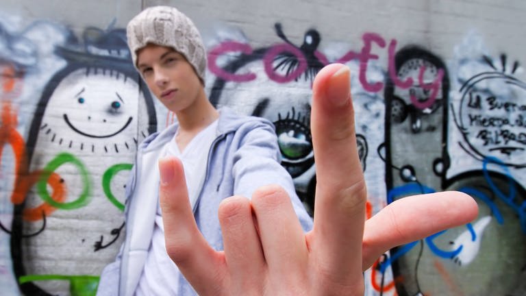 Cooler Jugendlicher mit Mütze vor Graffitiwand macht die Mano Cornuto, bei der man den Zeigefinger und den kleinen Finger nach oben streckt und die anderen Finger nach unten beugt. (Foto: IMAGO, IMAGO / blickwinkel)