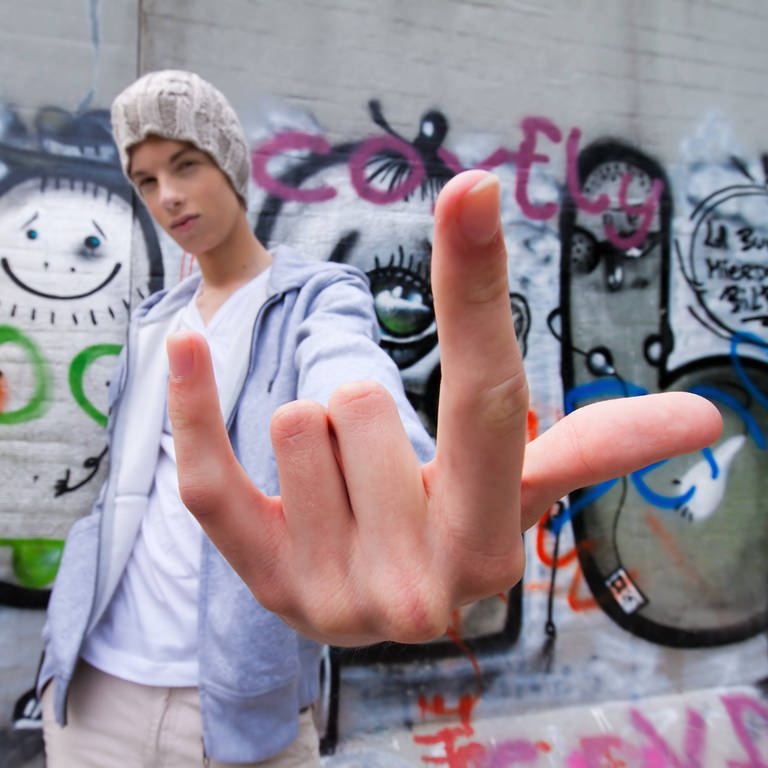 Cooler Jugendlicher mit Mütze vor Graffitiwand macht die Mano Cornuto, bei der man den Zeigefinger und den kleinen Finger nach oben streckt und die anderen Finger nach unten beugt.