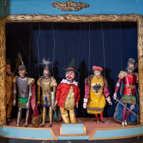 Ein historisches Puppentheater von circa 1870 steht im Münchner Marionettentheater neben der Bühne: Marionetten erscheinen auf der Bühne u. a. deshalb so groß, weil die Requisiten – also die Gegenstände, mit denen sie zu tun haben, wie Stühle, Tische oder Türen – im Verhältnis etwas kleiner gestaltet sind.