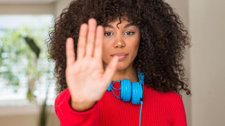 Junge Frau mit Kopfhörern signalisiert mit einer Handfläche "Stop": Triggerwarnungen kommen oft vor in Podcasts oder Videos, in denen Gewalt thematisiert wird. Sie sollen vor belastenden Inhalten schützen. Klappt das? (Foto: Getty Images, IMAGO / agefotostock)