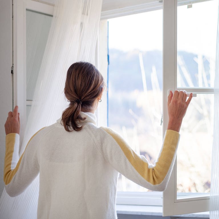 Eine Frau steht am Fenster und öffnet es weit mit Blick nach draußen: Richtiges Lüften hängt von den Umständen ab. Wie warm und feucht ist die Wohnung? Sind die Räume gut gedämmt? Trotzdem gibt es ein paar Grundregeln, die man beachten kann. (Foto: IMAGO, IMAGO / Roman Möbius)