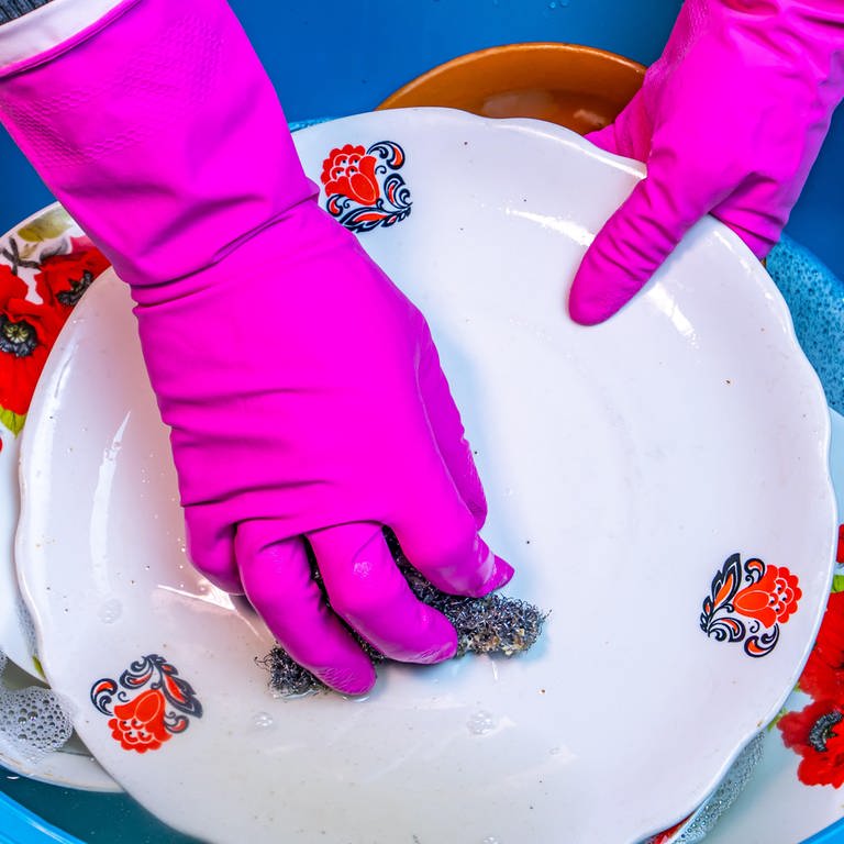 Hände in pinkfarbenen Handschuhen spülen einen Teller: Beim Spülen sollte man darauf achten, die Lappen regelmäßig zu wechseln. Spültücher mit eingewebten Silverfäden bringen keinen zusätzlichen Nutzen in Bezug auf die Hygiene.