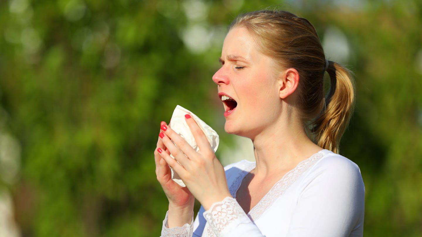 Eine junge Frau niest in der grünen Natur in ein Taschentuch: Niesen ist physiologisch ein explosives Atmen; die gleichen Körperpartien sind beteiligt wie beim normalen Atmen. Das 