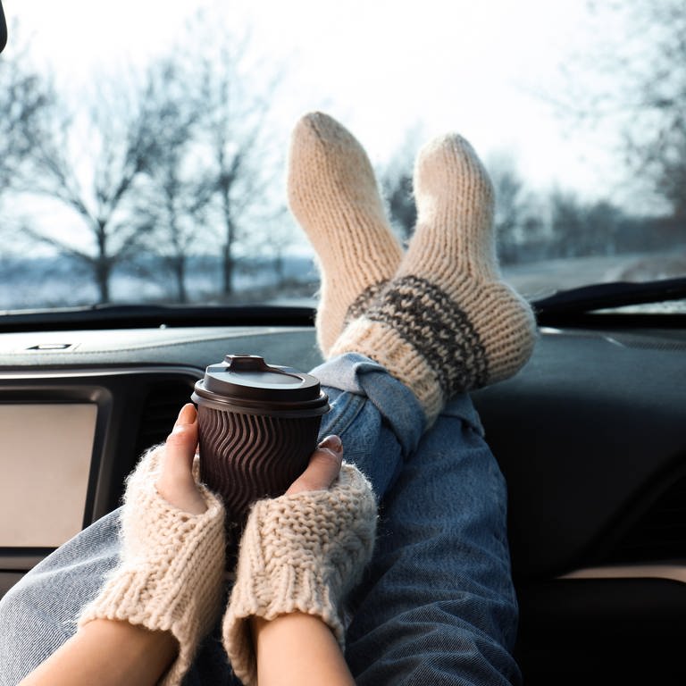 Junge Frau in warmen Socken hat ihre Beine aufs Armaturenbrett eines Autos gelegt und hält einen Becher Kaffee oder Tee: Tee und Kaffee sorgen für Harndrang und kalte Füße. Das liegt am Koffein. (Foto: IMAGO, IMAGO / Pond5 Images)
