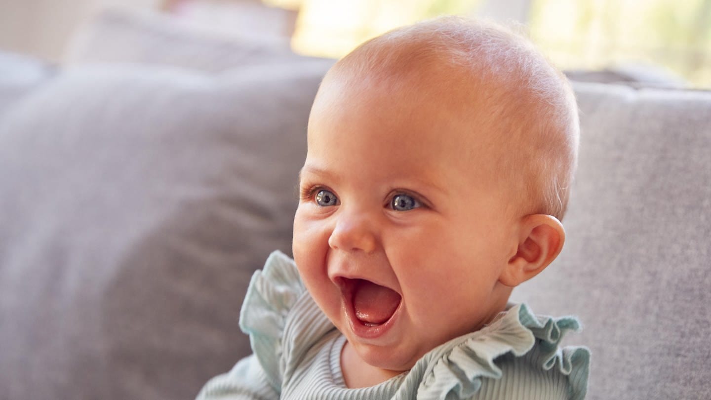 Säugling sitzt auf einer Couch und lacht:  Wenn ein Baby lacht, wenn man es kitzelt – und das tut es schon mit vier Monaten – ist das wirklich schon Humor? Oder nur ein Reflex oder einfach soziales Lachen? (Foto: picture-alliance / Reportdienste, picture alliance / Shotshop | Monkey Business 2)