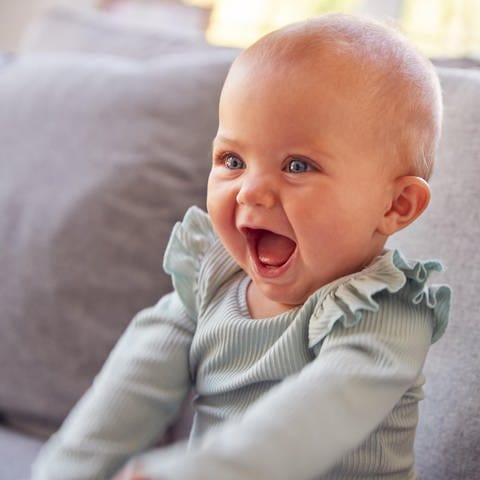 Säugling sitzt auf einer Couch und lacht:  Wenn ein Baby lacht, wenn man es kitzelt – und das tut es schon mit vier Monaten – ist das wirklich schon Humor? Oder nur ein Reflex oder einfach soziales Lachen? 