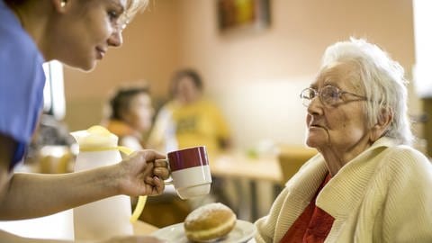 Pflegerin reicht einer alten Dame eine Tasse (Foto: IMAGO, imago/allOver-MEV)