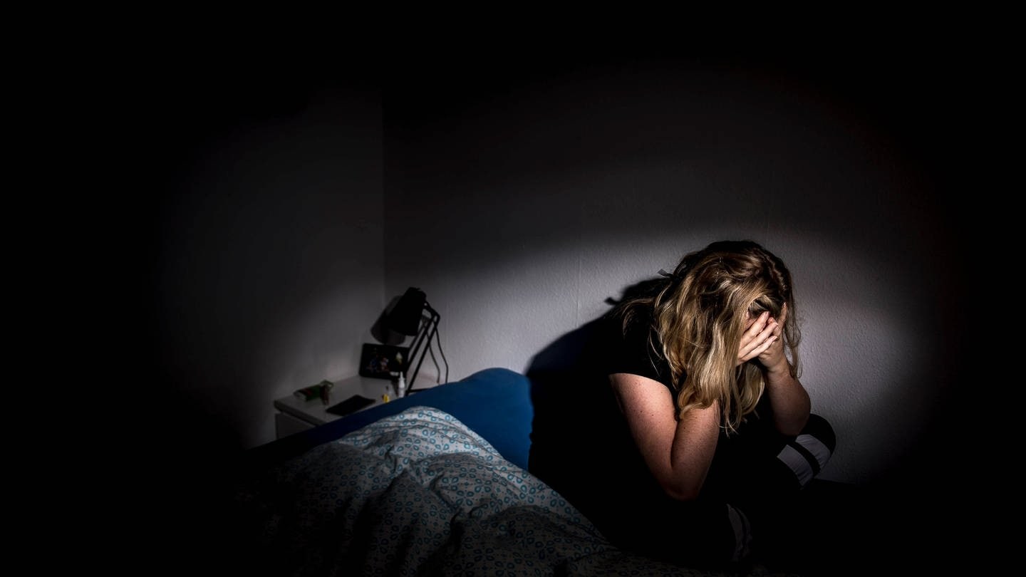 Eine Frau sitzt, das Gesicht in den Händen vergraben, in einem dunklen Raum auf einem Bett. (Foto: IMAGO, Imago Images / Ritzau Scanpix)