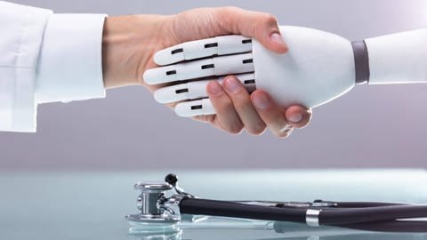 Ein Mensch und ein Roboter schütteln sich die Hände. Darunter liegt ein Stethoskop (Foto: IMAGO, imago images / Panthermedia)