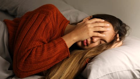 Junge Frau im Bett hält sich das Gesicht mit den Händen zu. (Foto: imago images, IMAGO / Panthermedia)