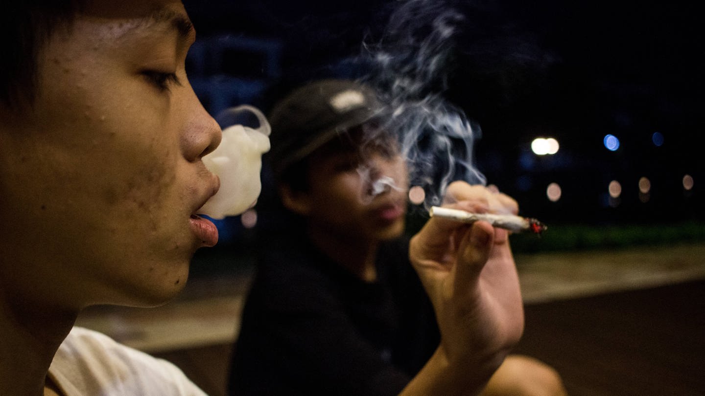 Jugendlicher atmet Rauch aus und hält einen glühenden Joint.