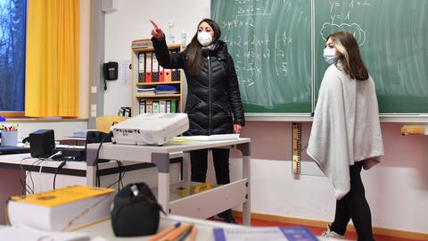 Lehrerin und Schülerin stehen an einer Tafel. Beide tragen Masken und da die Fenster geöffnet sind, haben sie dicke Jacken an. (Foto: IMAGO, IMAGO / Sven Simon)