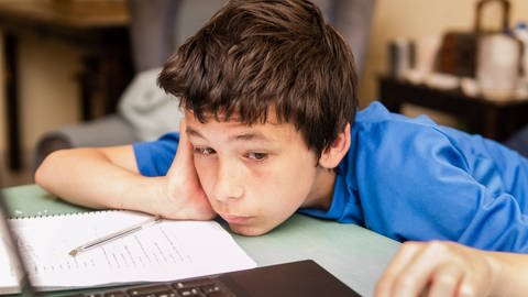 Frustriertes Kind lehnt sich an Schreibtisch mit Schreibutensilien und Laptop. (Foto: IMAGO, IMAGO / CHROMORANGE)