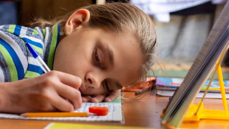 Kind lehnt kopf auf Schreibtisch und schreibt. (Foto: IMAGO, IMAGO / Shotshop)