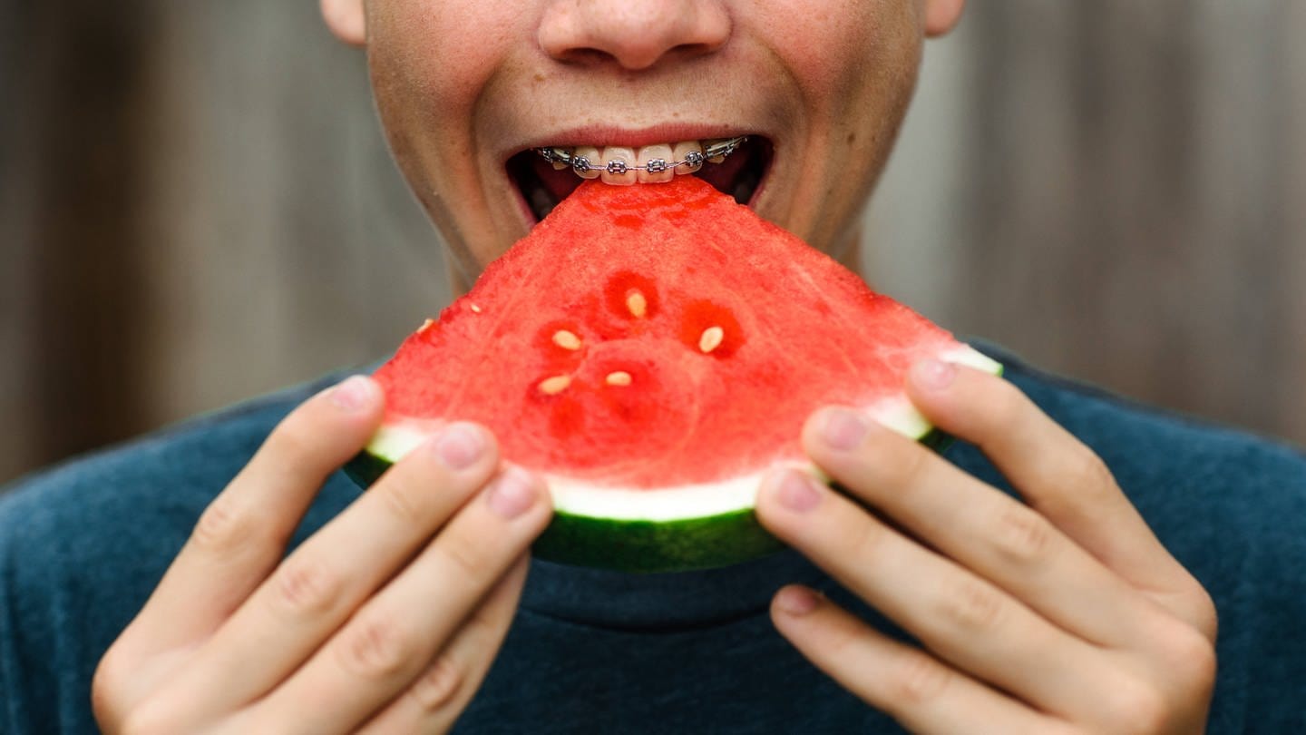 Nahaufnahme eines Munds mit Zahnspange, welcher in ein rotes Stück Melone beißt (Foto: IMAGO, Cavan Images via www.imago-images.de)
