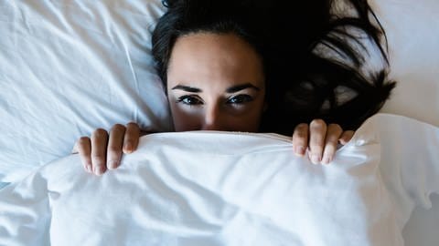 Frau hält sich die Bettdecke vor das Gesicht, ihre Augen sind geöffnet (Foto: IMAGO, Cavan Images via www.imago-images.de)