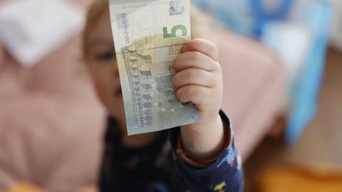 Kind hält 5-Euroschein in der Hand: Das Kindergeld kommt bei Alleinerziehenden und vielen Familien, die Hartz IV beziehen, fast nicht an, weil es verrechnet wird (Foto: IMAGO, IMAGO / photothek)