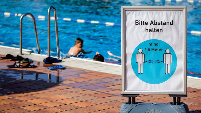 Ein Schild mit der Aufschrift "Bitte Abstand halten" steht vor dem Schwimmbecken eines Freibads.