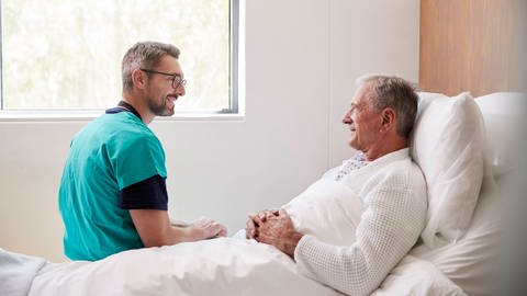 Arzthelfer im Gespräch mit einem Patienten, der in einem Krankenhausbett liegt (Foto: IMAGO, imago images/Shotshop)