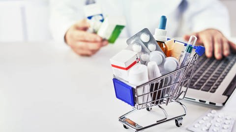 Kleiner Einkaufswagen mit zahlreichen Medikamenten, im Hintergrund eine Person mit Laptop und Gesundheitskarte (Foto: IMAGO, xfoodandmorex Panthermedia25935172)