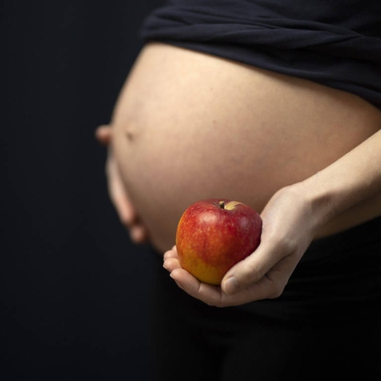 Ernährung während der Schwangerschaft: Frau hält einen Apfel vor Babybauch (Foto: IMAGO, IMAGO / photothek)