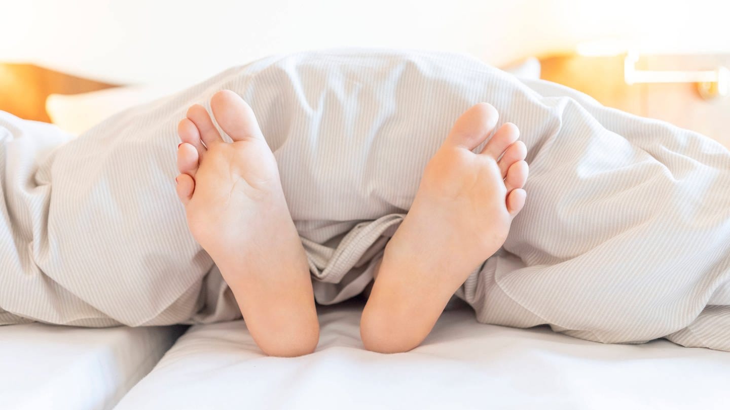 Zwei Füße schauen unter einer Bettdecke hervor (Foto: IMAGO, IMAGO / Westend61)