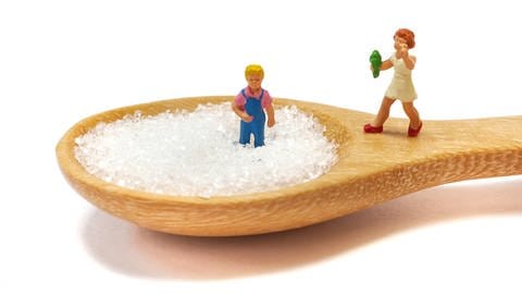 Zwei Kinderspielzeugfiguren stehen auf einem Holzlöffel voller Zucker. (Foto: IMAGO, IMAGO / agefotostock)