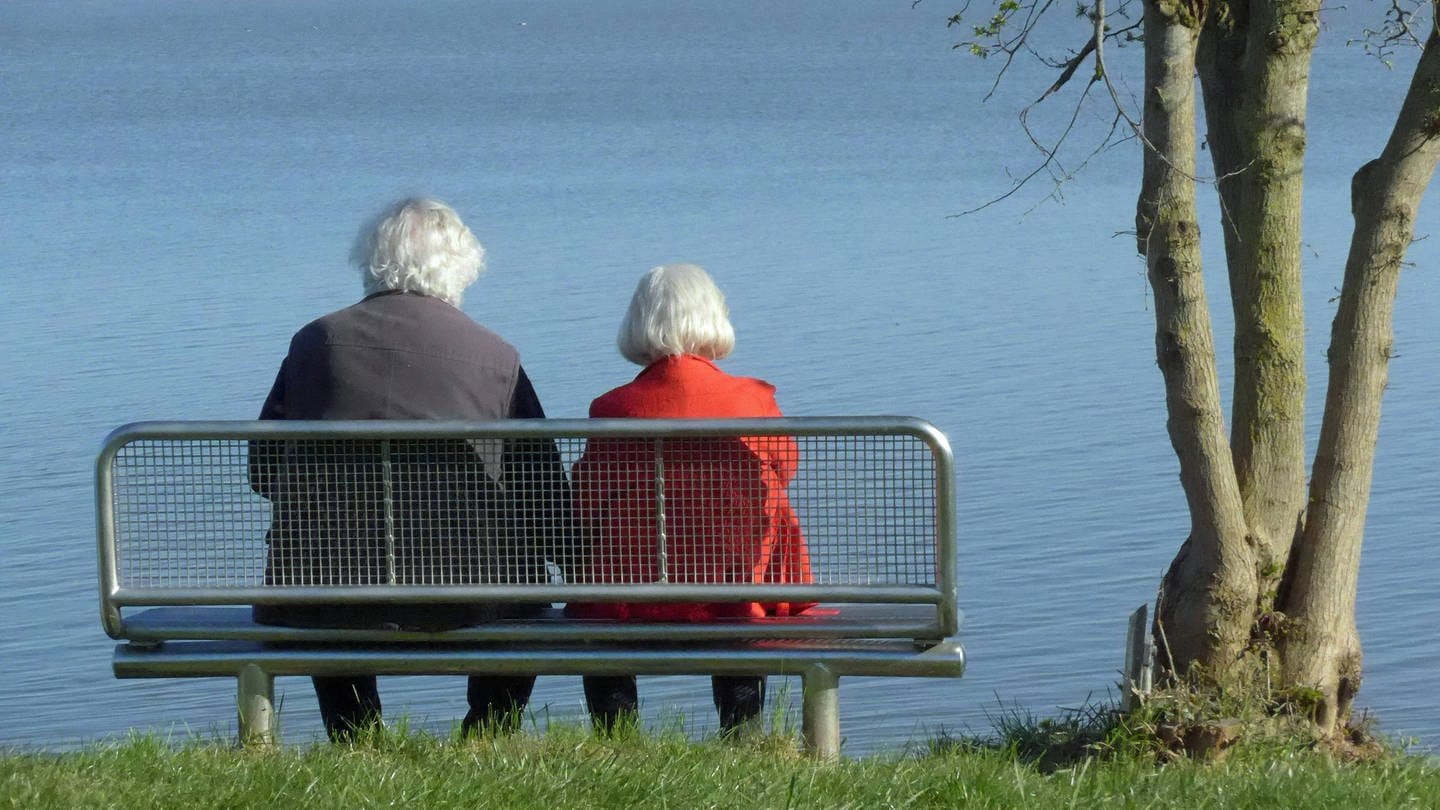 Ein Seniorenehepaar sitzt auf einer Bank am See und schaut aufs Wasser. (Foto: IMAGO, IMAGO / Eckhard Stengel)