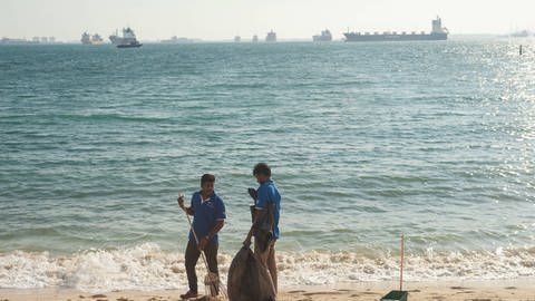 Zwei Arbeiter säubern den Strand während im Hintergrund zahlreiche Schiffe am Horizont fahren. (Foto: IMAGO, imago images/Olaf Schuelke)